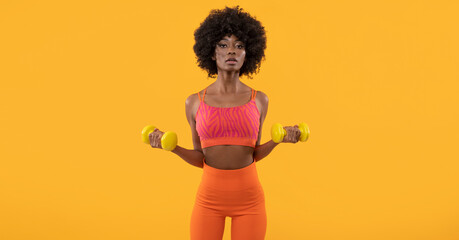  Dark skin female fitness trainer with afro hair and dumbbells on isolated orange background.