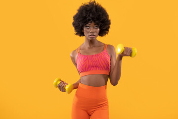  Dark skin female fitness trainer with afro hair and dumbbells on isolated orange background.