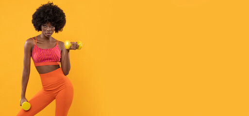  Dark skin female fitness trainer with afro hair and dumbbells on isolated orange background.