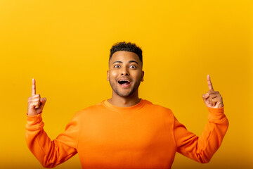 Attractive indian man with astonished shocked facial expression pointing up with both fingers, presenting copy space. Indoor studio shot isolated on yellow background
