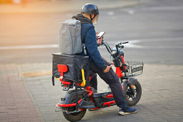 Naklejka premium Online delivery service, delivering food with e-scooter. Courier rides electric moto bike, deliver food orders to customers. Boy in helmet ride electric scooter with thermal box, fast food delivery.