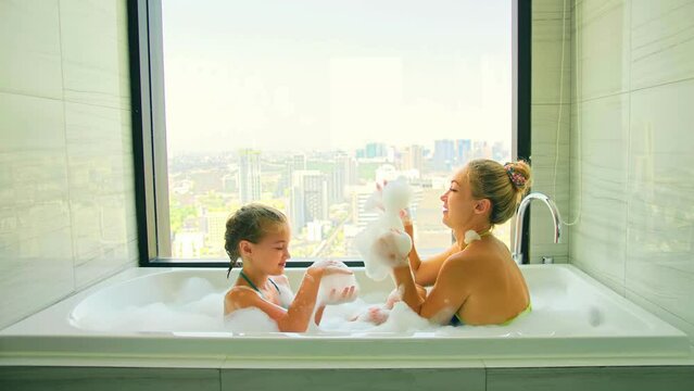 Funny mother and daughter bonding activity, play with foam, laughing and smiling in bath with stunning city. Cheerful mother and daughter having fun in bathroom.Cute mom and child take bath with foam