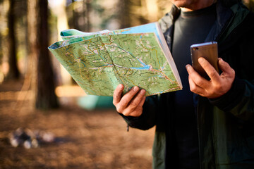 Adult hiker with backpack using mobile phone and map in forest near his tent. Hiking elderly man in autumn nature holding map outdoors next to a campsite in the sunlight closeup shot. Ageless travel