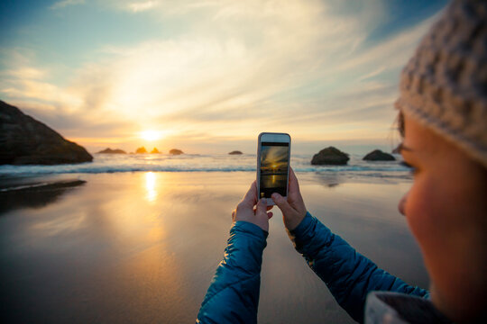 A young woman uses her smartphone to take a picture of the sunset from Bandon Beach, Oregon.