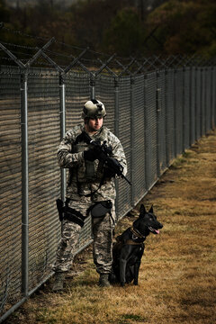 An Air Force Security Forces K-9 Handler, And His Military Working Dog, Stand Next To A Barbed Wire Fence.