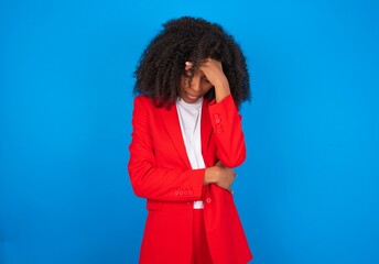 young businesswoman with afro hairstyle wearing red over blue background making facepalm gesture...