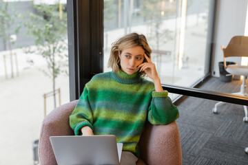 thoughtful young woman in sweater sitting with laptop in lobby of hotel in Barcelona.