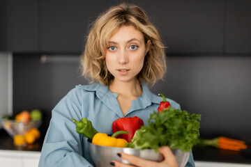 blonde young woman with wavy hair holding bowl with organic vegetables in kitchen.