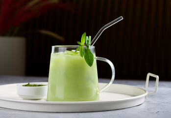 Matcha latte served on a tray on restaurant table with textured interior background. Transparent cup and a straw with organic drink of ground powder of green tea leaves with milk, ice cubes and mint. 