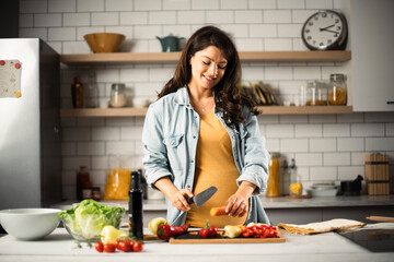 Young woman cutting vegetables in kitchen. Beautiful pregnant woman making salad.