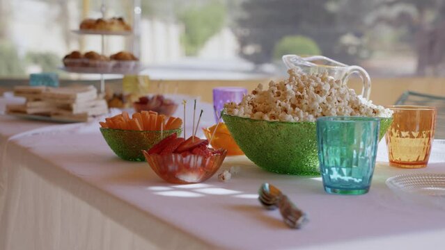 Approaching to a table with sweets for a party in summer, with a tray with croissants, sandwich, popcorn, crudit&eacute;s, coloured glasses and white tablecloth moving blowed by the wind.
