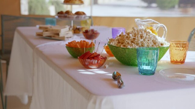 Table with sweets for a party in summer, with a tray with croissants, sandwich, popcorn, crudit&eacute;s, coloured glasses and white tablecloth moving blowed by the wind.