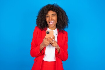 Pleased young businesswoman with afro hairstyle wearing red over blue background using self phone and looking and winking at the camera. Flirt and coquettish concept.