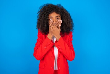 Stunned young businesswoman with afro hairstyle wearing red over blue background covers mouth with both hands being afraid from something or after hearing stunning gossips.