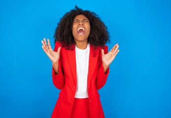 Crazy outraged young businesswoman with afro hairstyle wearing red over blue background screams loudly and gestures angrily yells furiously. Negative human emotions feelings concept