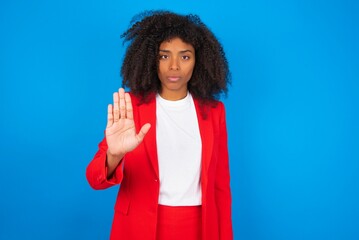 young businesswoman with afro hairstyle wearing red over blue background shows stop sign prohibition symbol keeps palm forward to camera with strict expression