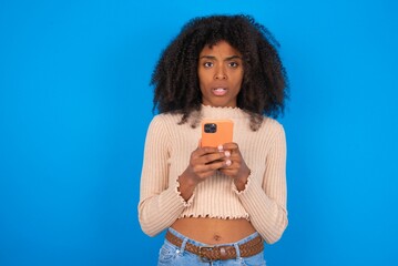 Photo of astonished Young woman with afro hair style wearing crop top over blue background hold smartphone dislike feedback concept