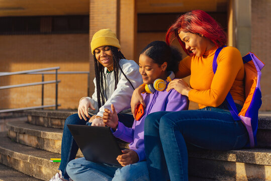 Three Hispanic Girls Sitting On The Stairs Of The College Campus Working On A Laptop. Latina Friends And Classmates Outside The College Using Technology.