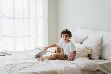 Handsome curly caucasian baby boy sitting on bed holds tablet looks at camera on sunny day. Childhood and gadgets. Smart Hispanic little kid enjoys games via internet. Cute toddler plays home.