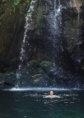 Young woman swimming at lake under small waterfall in Isalo park jungle, water stream falling over large rocks