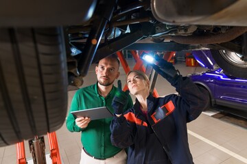 A service station manager and a young female car mechanic are standing under a car on a lift. Diagnostics of the suspension and steering of the car