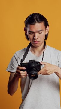 Vertical video: Young professional photographer using dslr camera to take pictures, having fun with photos at photography shoot in studio. Male model posing with equipment, feeling confident.