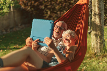 happy senior couple relaxing on hammock - laughing positive retirees watching video on tablet