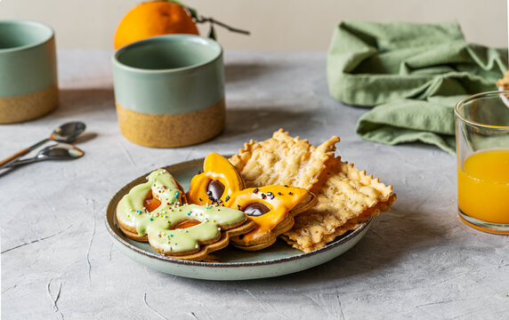Carnival sweets in shape of carnival mask green and orange colour, mugs, orange fruit and juice, carnival fritters or bugie on light grey background