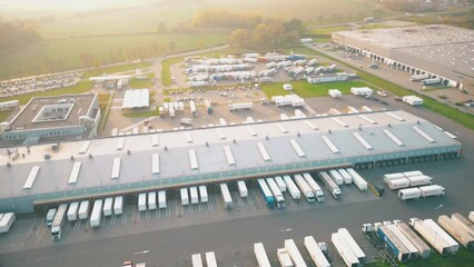 Logistics park with a warehouse and loading hub. Semi-trucks with cargo trailers standing at the ramps for loading/unloading goods at sunset. Aerial hyper lapse.