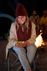 Front view of Caucasian woman sitting outside in a camping using phone to be connected and sending messages happy, while friends in the background enjoying the bonfire. Vertical photo.