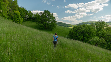 Fototapeta na wymiar A young person is walking on a meadow in a green hill landscape with trees and grass. The sky above is blue with soft sheep clouds
