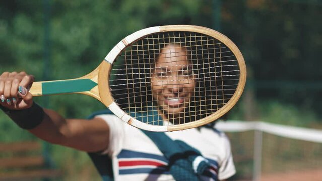 4k African american woman posing with retro vintage tennis racket on clay tennis court in sunny day. Woman playing tennis in vintage sportwear.