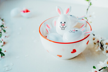 Close up Funny Easter eggs containers with bunny ears filled with candy and chocolate in a bowl on the white kitchen table with blooming branches. Festive spring Easter composition. Selective focus