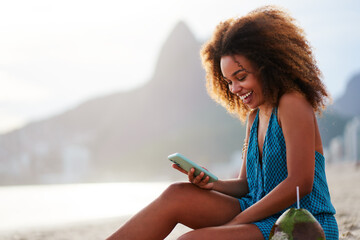 portrait smiling happy young brazilian woman sitting on the beach holding and looking at cell phone...