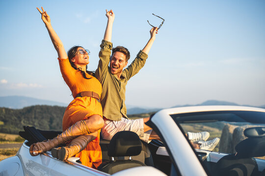 Sunset sun bath for a couple of lovers on the go, travel and holiday lifestyle. young couple having fun standing on convertible car, countryside background