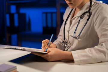 Unknown female doctor sitting in clinic and using clipboard during night shift. Perfect medical service in hospital. Medicine and healthcare concept