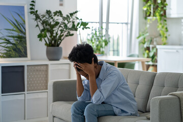 Hopeless African American female suffering from of wrong decision crying sitting on sofa alone at...
