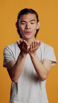 Vertical video: Male model blowing air kisses in studio, acting silly and sensual on orange backdrop. Carefree man sending kisses and being flirty, acting romantic with kissy funny face. Natural