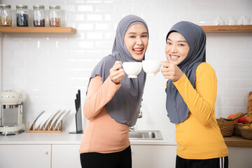 Cheerful Muslim friends drinking coffee standing in front of kitchen at home. Arabic Young women beautiful and bonding life spending time together on weekend indoors.