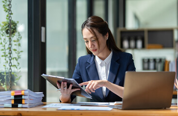 Beautiful attractive Asian businesswoman holding a clipboard of documents sitting analytically analyzing various marketing management information the finances were at her desk in the office at ease. © crizzystudio