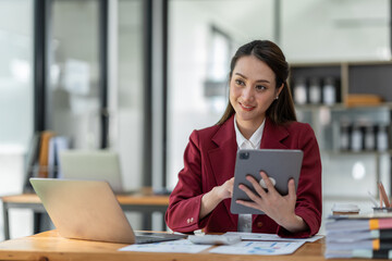 Asian businesswoman in a suit sitting holding iPad or tablet in contact See details and discuss business information happily in the office.