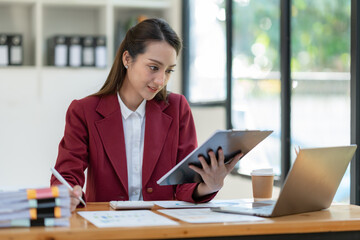 Beautiful attractive Asian businesswoman holding a clipboard of documents sitting analytically analyzing various marketing management information the finances were at her desk in the office at ease. © crizzystudio