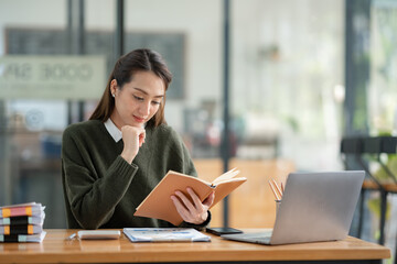 Fototapeta premium Beautiful Asian businesswoman working on a laptop and having fun at work and reading review book documenting detailed financial information earning peace of mind.