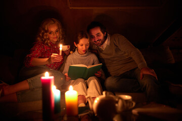 Cozy family evening. Mother, father and little girl sitting on sofa without electricity and reading...