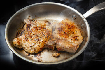 Two pork chops are roasted in a stainless steel frying pan on a black cooktop, food and cooking concept, selected focus
