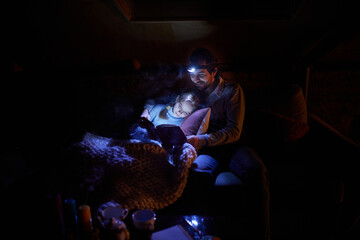 Father with his little daughter sitting on sofa at home in the evening and reading book with forehead battery lamp. No electricity, blackout. Leisure time. Concept of power outage, adjusting