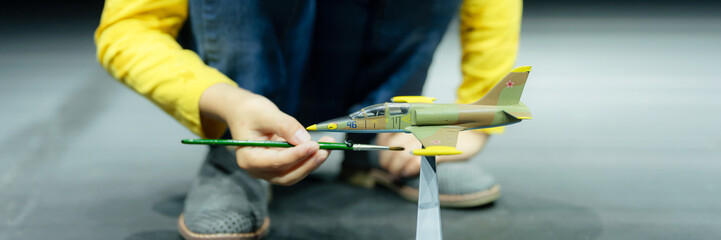 candid little six year old kid boy painting or colorize or colour model airplane with gouache paints while squatting on floor studio illuminated by light. banner