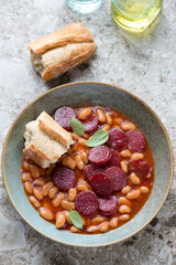 Stew with chorizo sausage and white beans served with crusty bread on a light-brown granite background, vertical shot, elevated view