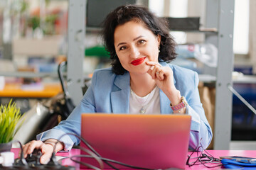 a brunette woman in a blue jacket sits at a desk and works on a laptop.
