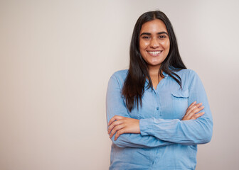 Young Indian woman stands arms folded beige wall, blue shirt office attire